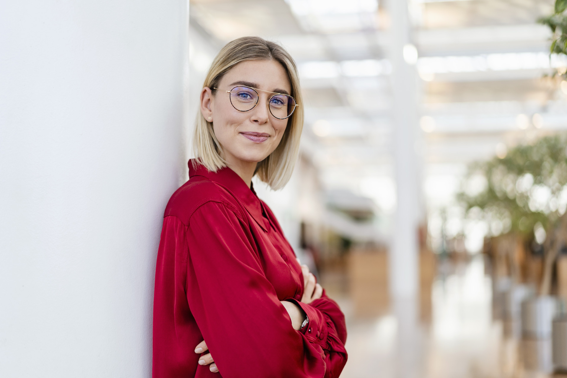 Frau mit Brille und blondem Haar und roter Bluse lehnt sich an Wand an. Hat die Arme verschränkt.