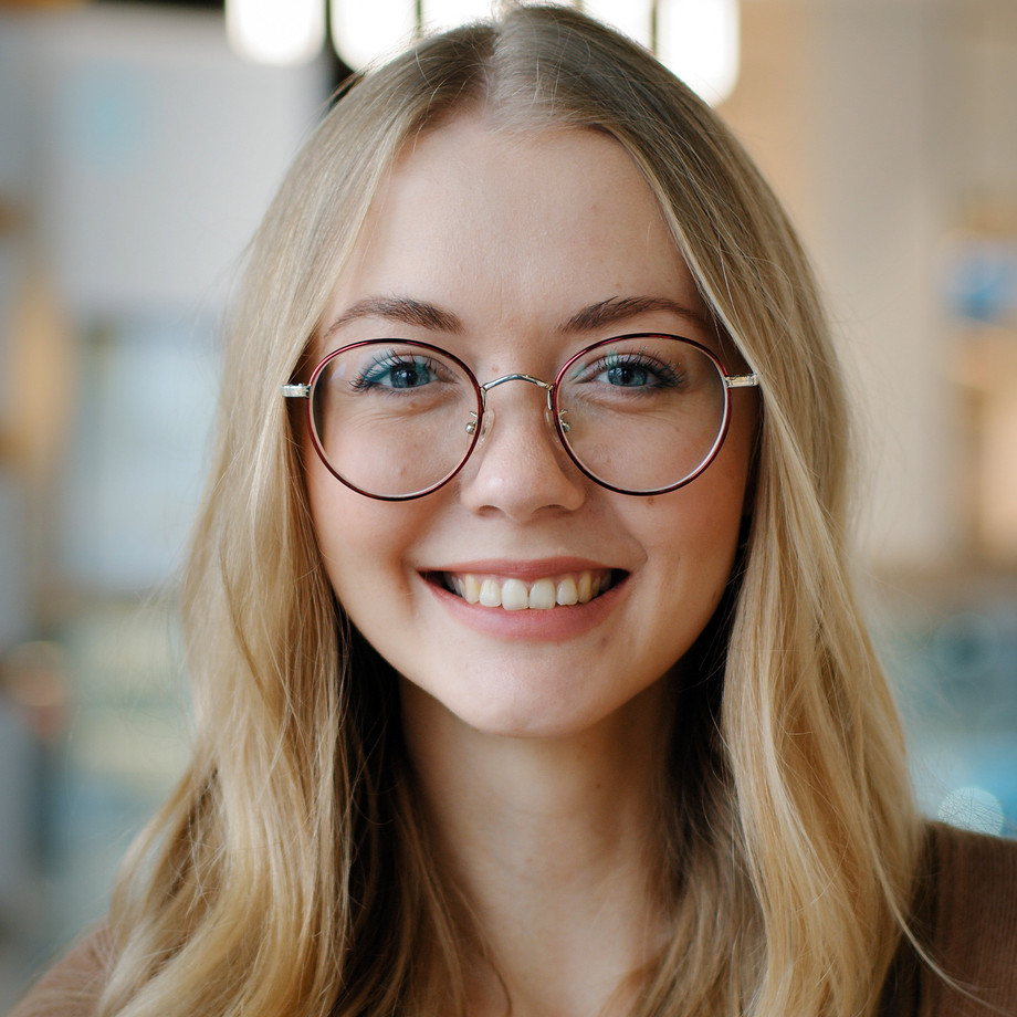 Junge Frau mit großer Brille und blondem Haar in einem Eingangsbereich einer Unternehmung